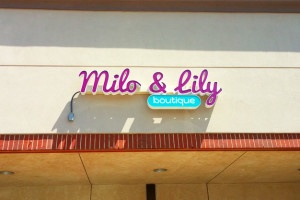 Picture of channel letter wall sign for Milo and Lily. The sun is bright, and the letters are dark purple, with an aqua colored capsule underneath that says "boutique."